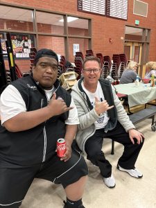 Two men sit at a table in a brick-walled room, smiling and giving thumbs up; one holds a Coca‑Cola can in his hand.